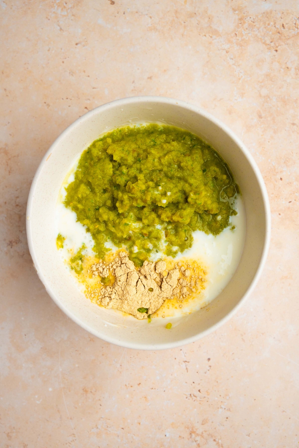 A white bowl with ingredients to make Chuy's jalapeno dip including mayo, milk, buttermilk, seasonings, and purred jalapeno mixture. The bowl is on a tan counter.