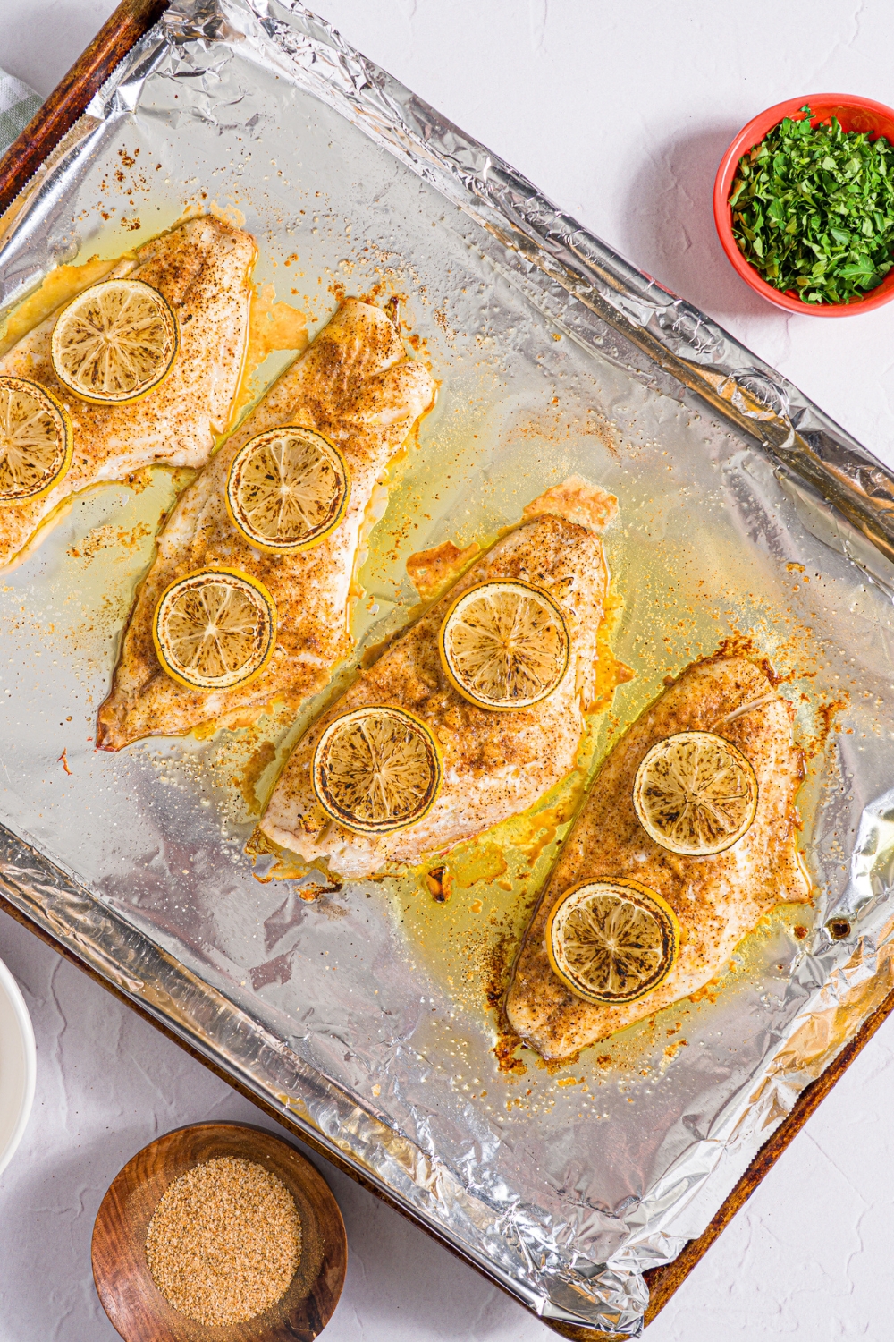 A baking sheet lined with foil with baked red snapper with a garlic topping and sliced lemon. The sheet is on a white counter with small bowls of garnishes.