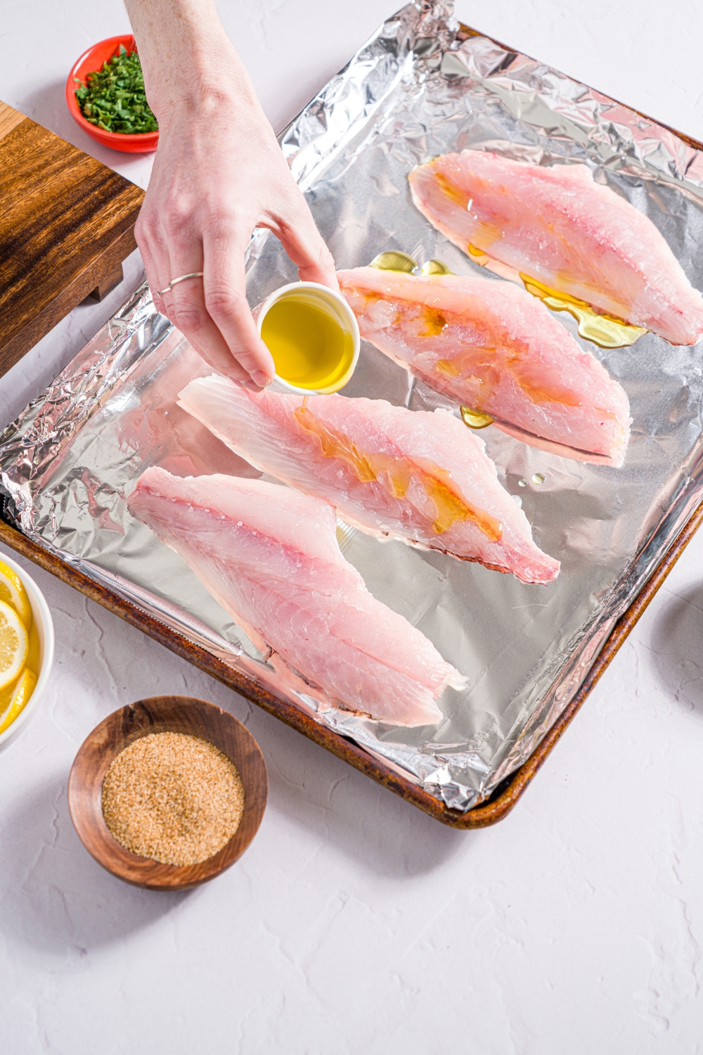 A baking sheet lined with foil with baked red snapper with a hand pouring olive oil over the fish. The sheet is on a white counter with small bowls of garnishes.