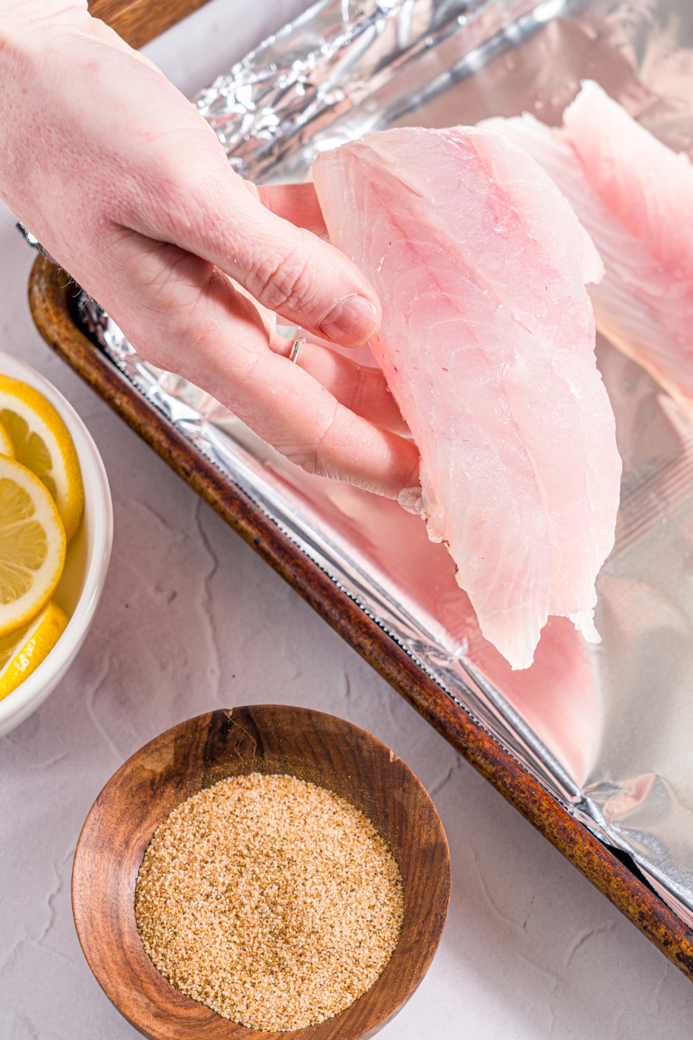 A hand placing a red snapper fillet on a baking sheet lined with foil. The sheet is on a white counter with a small bowl of ingredients.