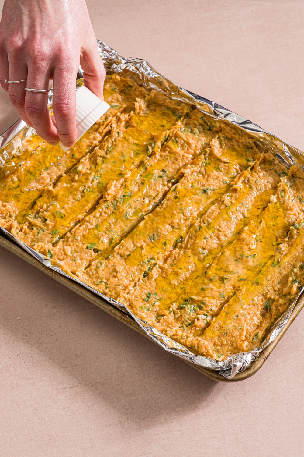 A baking sheet lined with foil with uncooked chicken kefta mixture spread on the sheet. A hand is pouring a small ramekin of oil over the mixture. The sheet is on a tan counter.