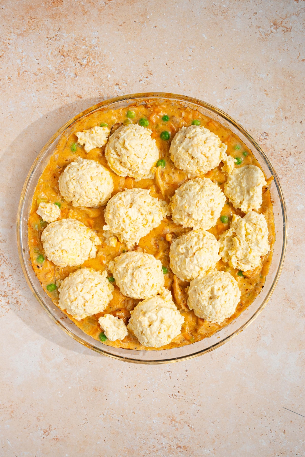 A round baking dish with chicken pot pie filling topped with uncooked cheddar bay biscuit dough. The dish is on a tan counter.