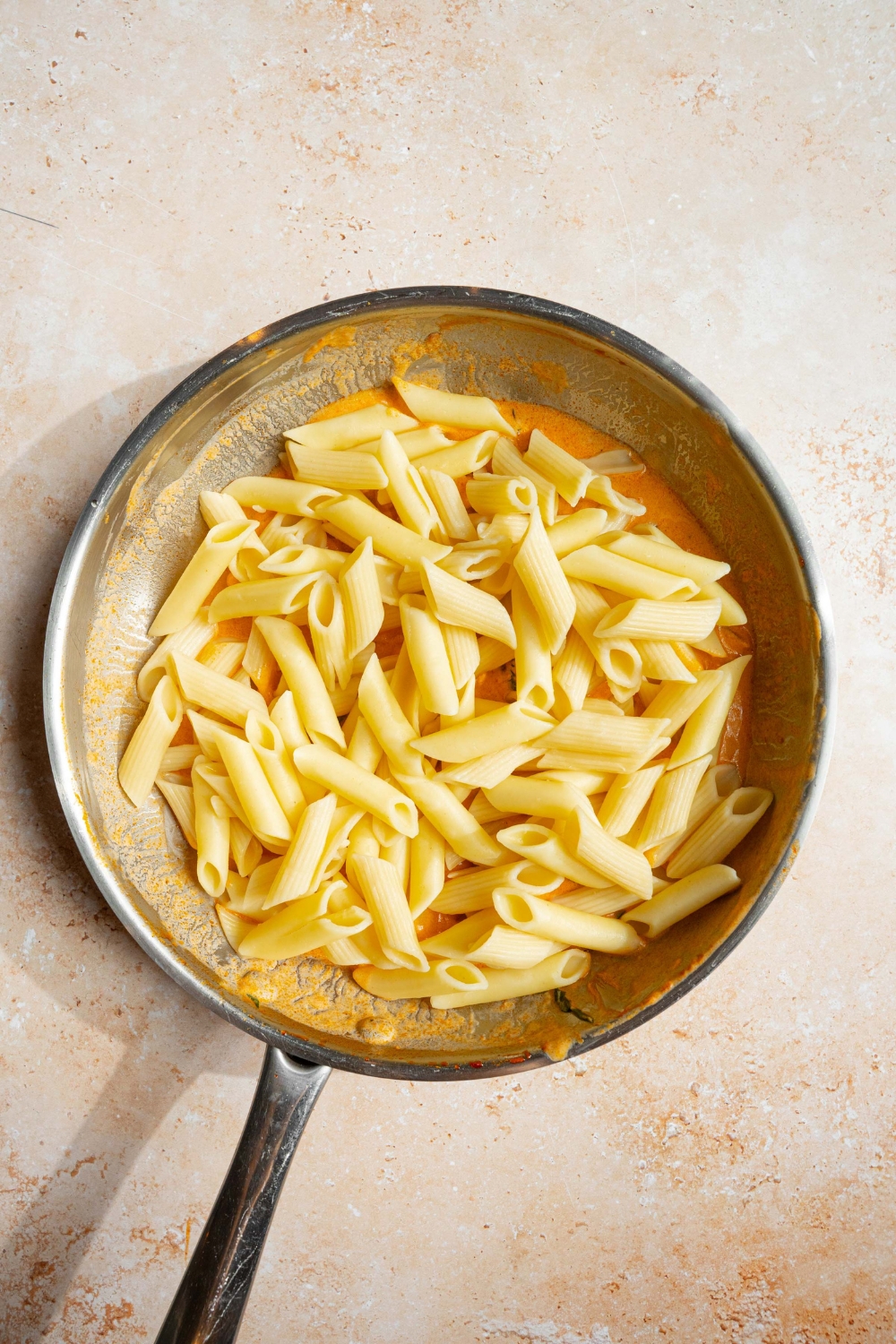 A skillet with blush sauce topped with penne pasta. The skillet is on a tan counter.