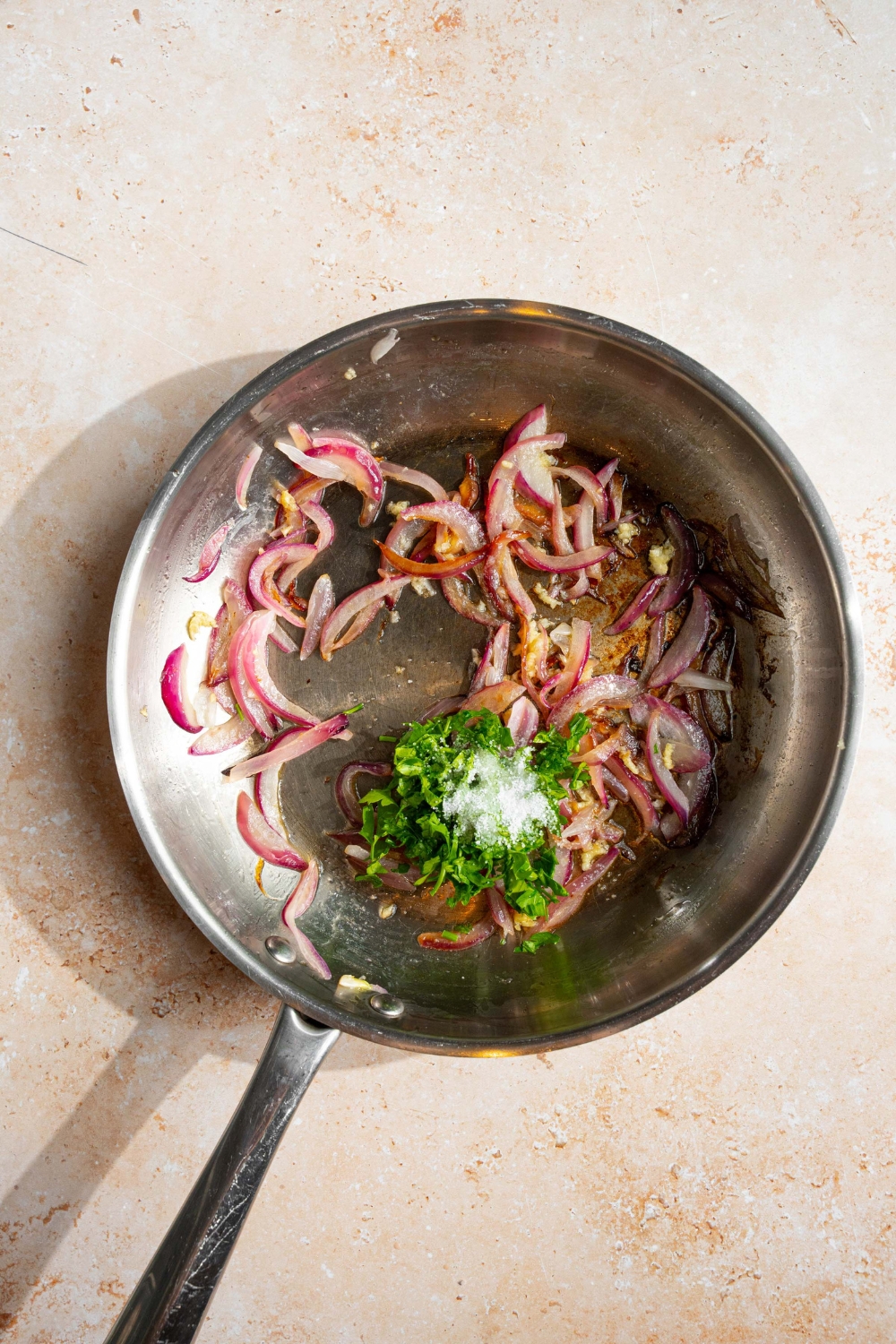 A skillet with sautéed sliced onion, garlic, and fresh parsley cooking in butter. The skillet is on a tan counter.