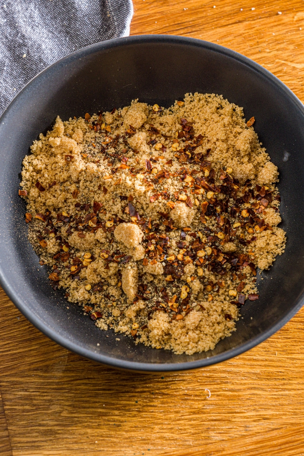 A black bowl with a brown sugar mixure with black pepper and red pepper flakes. The bowl is on a wooden counter with a blue cloth napkin.