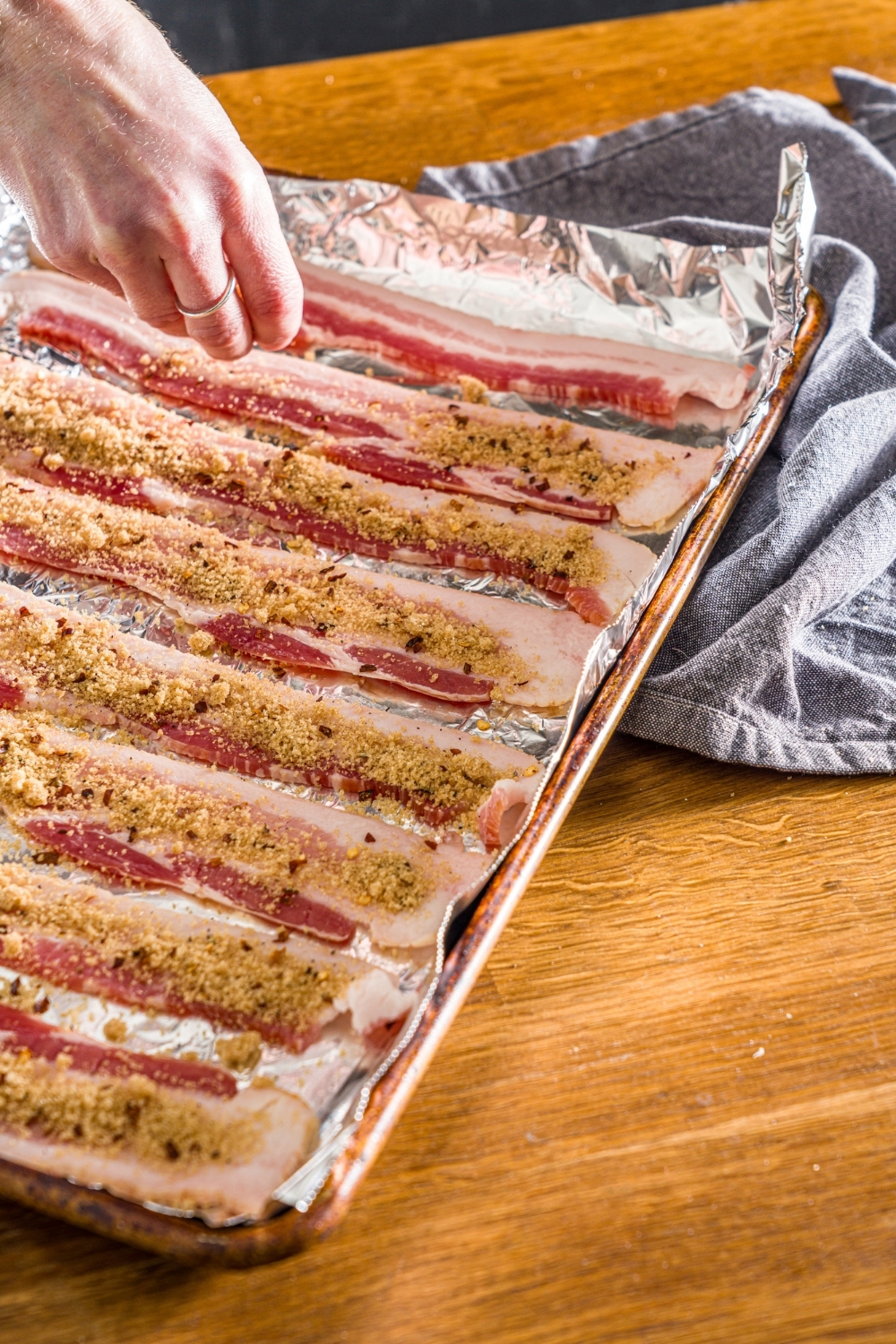 A baking sheet lined with foil with uncooked bacon being sprinkled with brown sugar mixture. The sheet is on a wooden counter with a blue cloth napkin.
