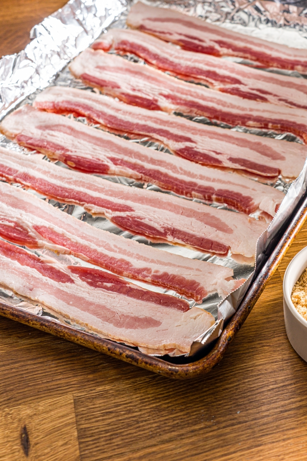 A baking sheet lined with foil with uncooked bacon. The sheet is on a wooden counter.
