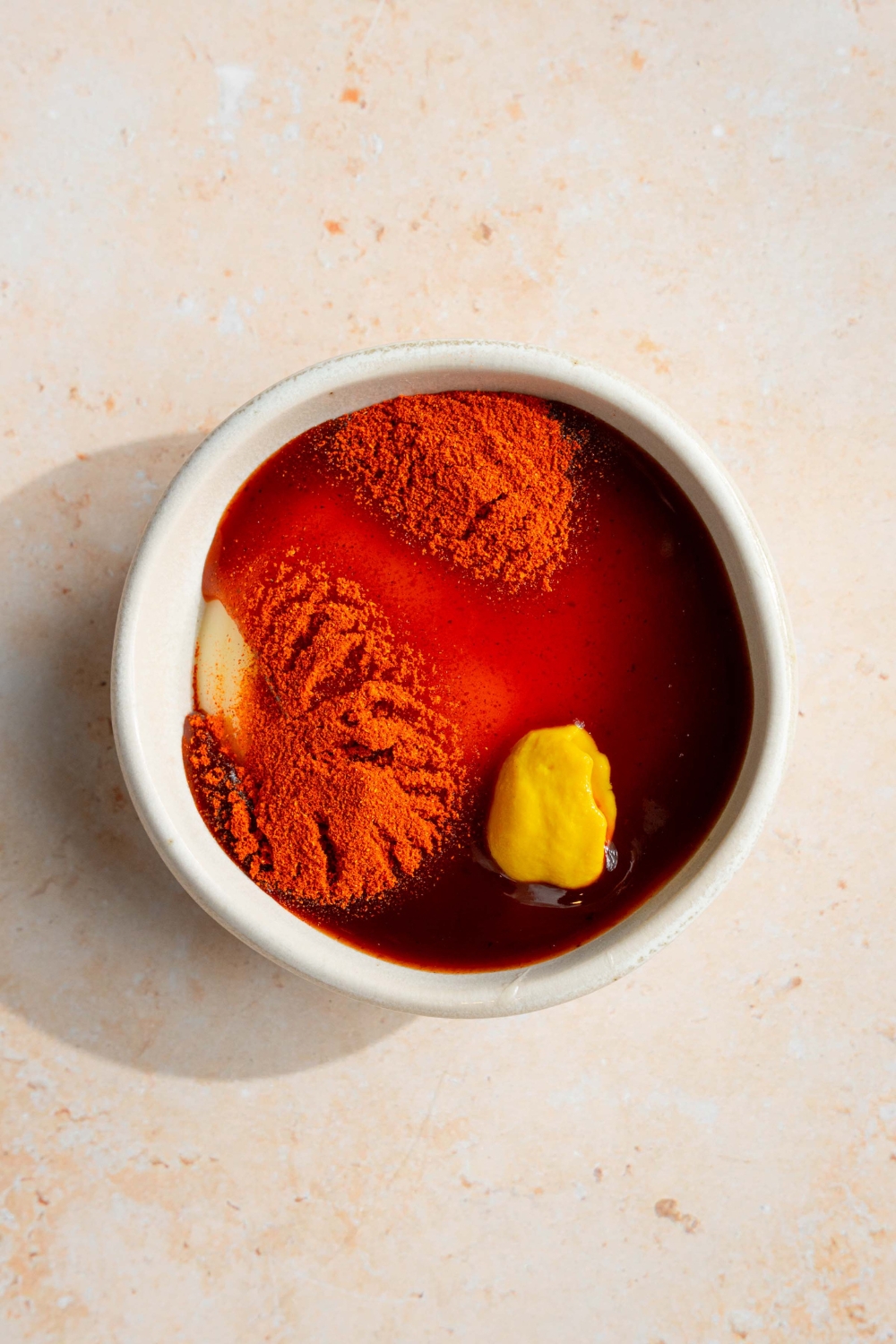 A white bowl with ingredients to make campfire sauce including mayo, BBQ sauce, brown mustard, and seasonings. The bowl is on a tan counter.