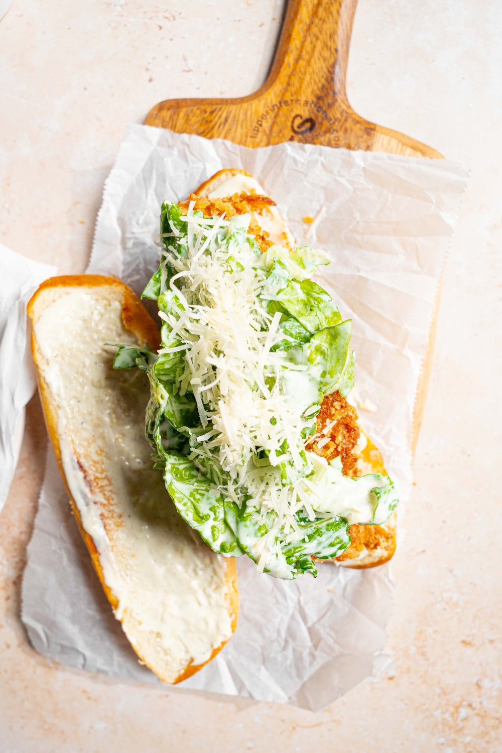 An open chicken caesar sandwich on a wooden board lined with parchment paper. The board is on a tan counter.