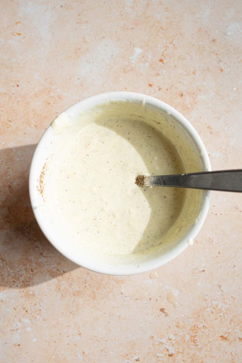 A white bowl with a spoon mixing caesar dressing. The bowl is on a tan counter.