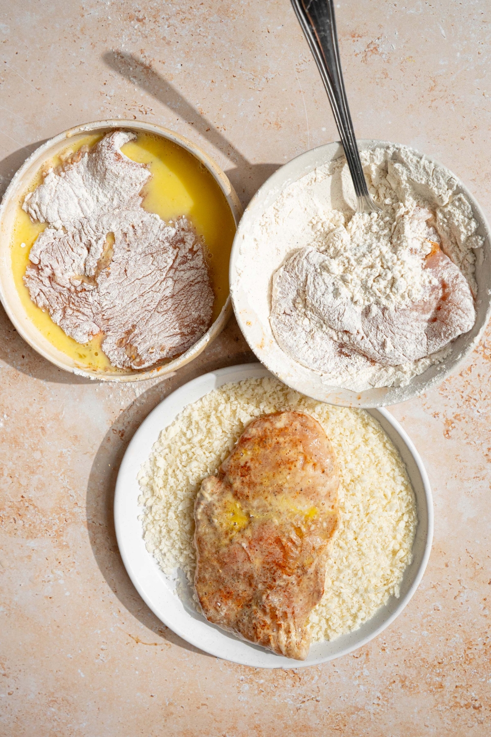 Three bowls with egg wash, flour mixture, and panko breadcrumbs, with a seasoned chicken breast dredged in each bowl. The bowls are on a tan counter.