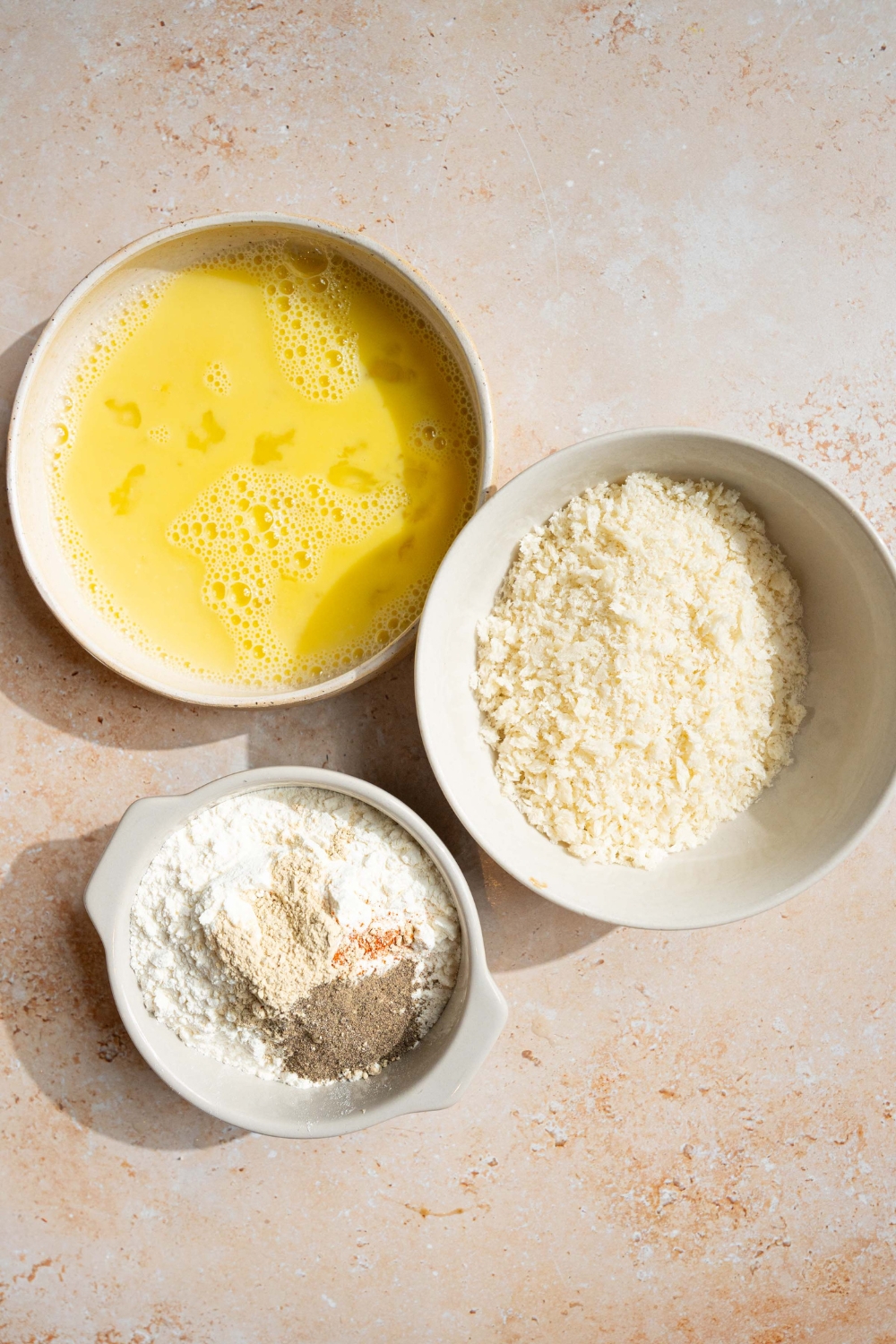 A white bowl of whisked egg, a white bowl with panko breadcrumbs, and a white bowl with a flour mixture.