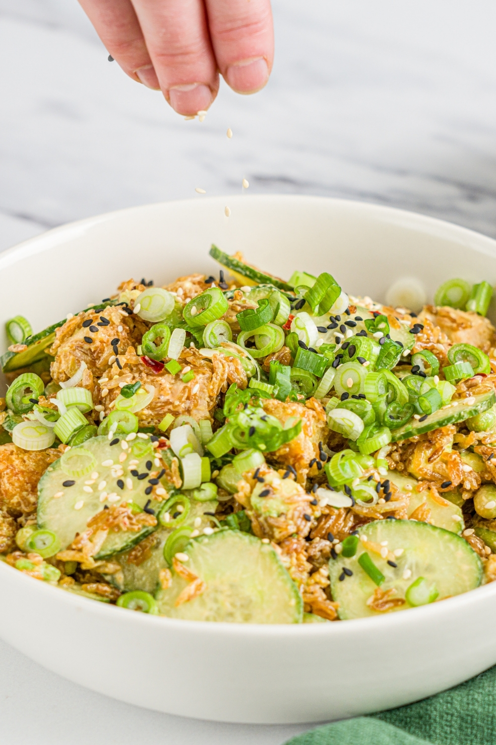 A white bowl with crispy rice chicken and cucumber salad garnished with sliced green onion. A hand is sprinkling sesame seeds over the salad. The bowl is on a marble counter.