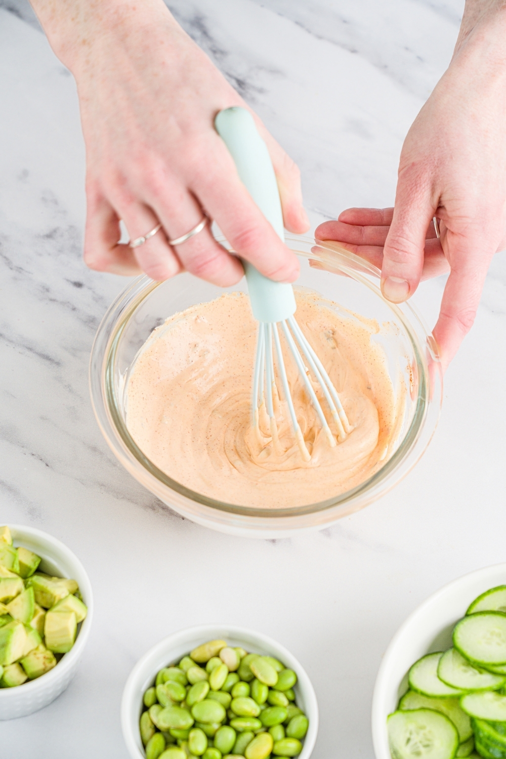A small glass bowl with a whisk mixing a mayo and sweet chili sauce dressing. The bowl is on a marble counter with a bowl of edamame, cucumbers, and avocado.