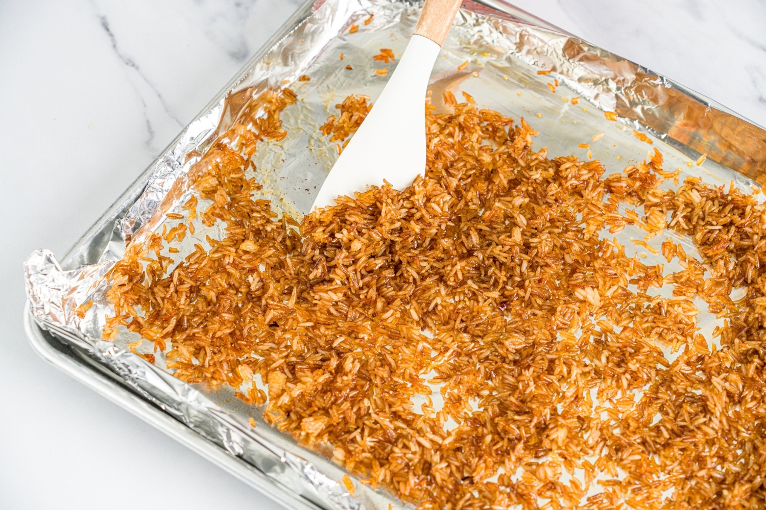 A baking sheet lined with aluminum foil with crispy rice. A spatula is mixing the rice. The sheet is on a marble counter.