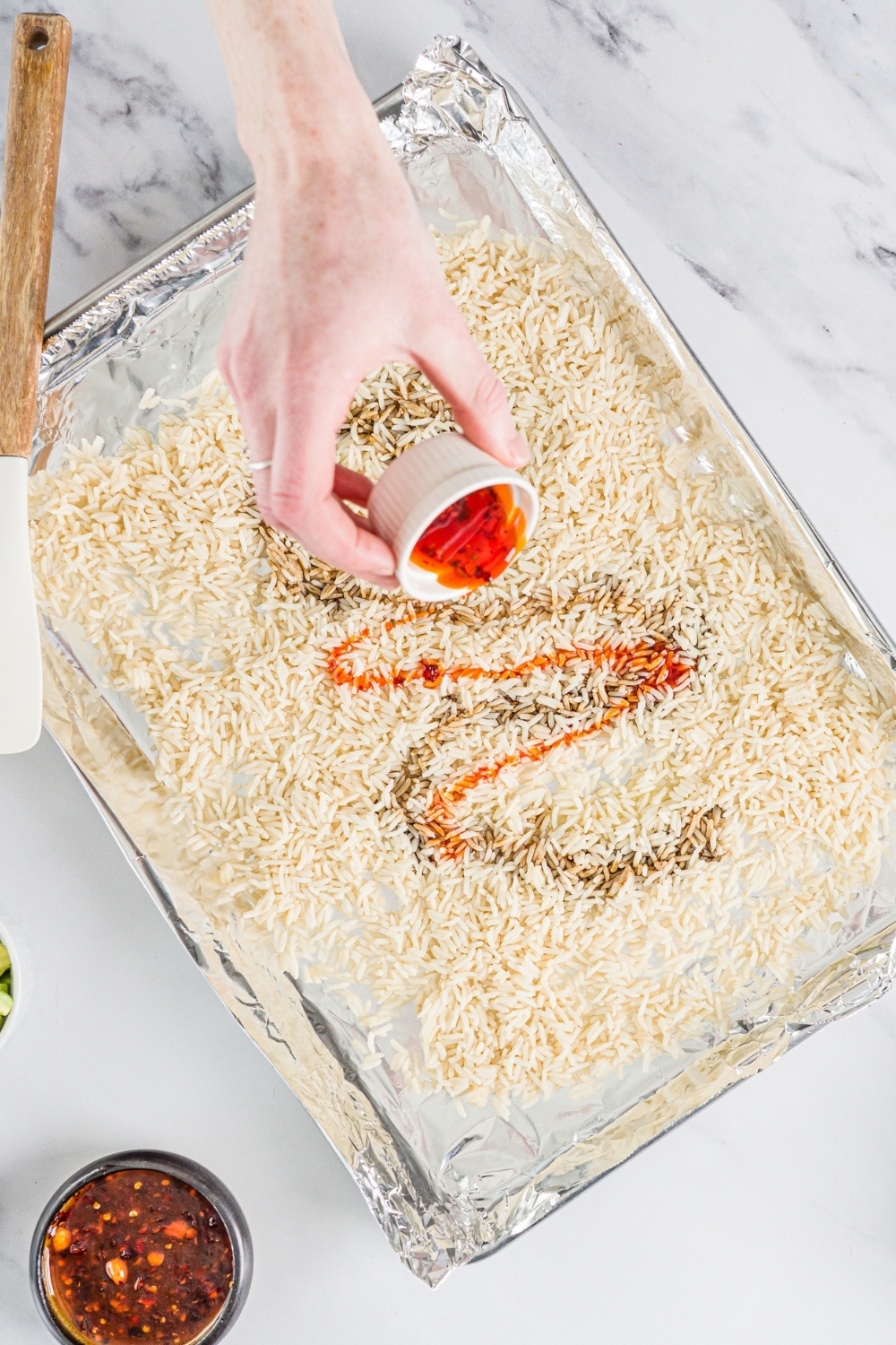 A baking sheet lined with aluminum foil with cooked rice. A hand is pouring a small bowl of chili soy sauce over the rice. The sheet is on a marble counter.