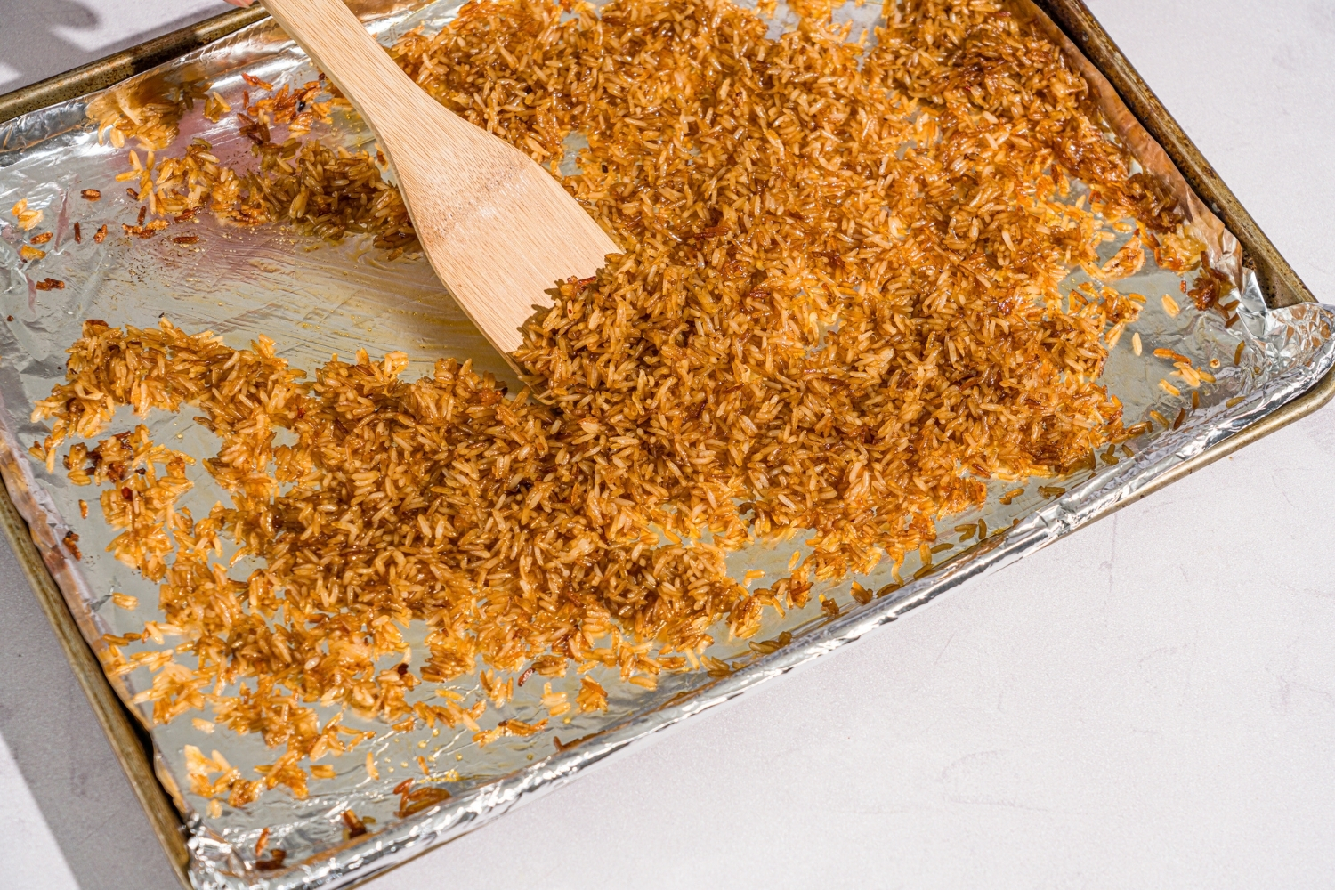 A baking sheet lined with foil with cooked rice. A spatula is spreading the rice around. The sheet is on a marble counter.