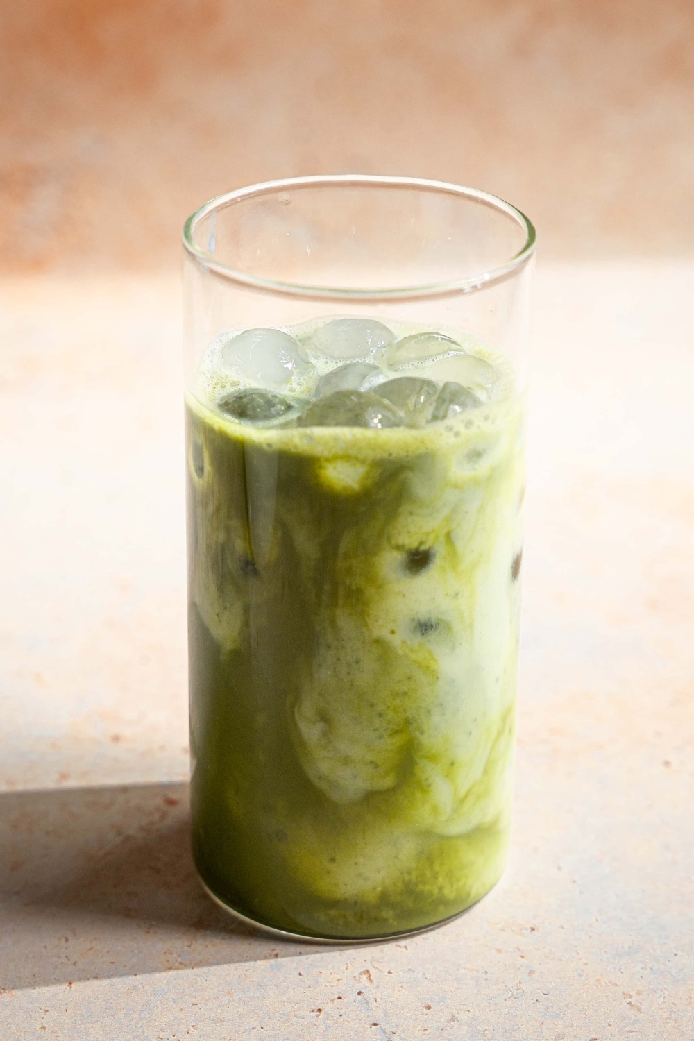 A tall glass with matcha tea and milk over ice. The glass is on a tan counter.