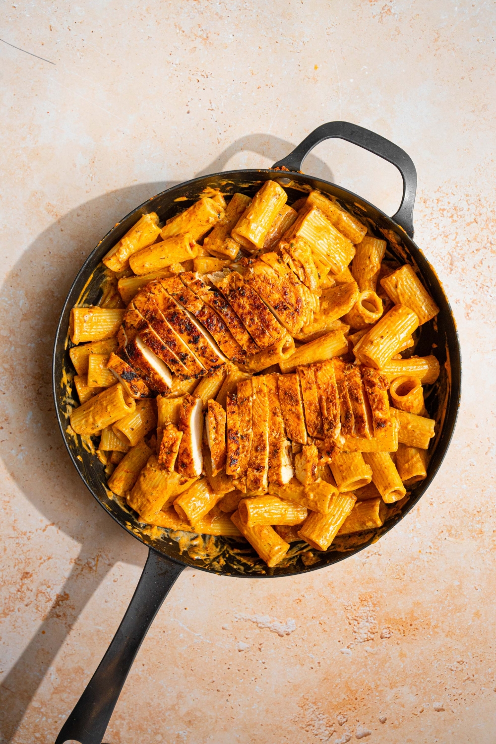 A skillet with garlic parmesan pasta with sliced chicken on top. The skillet is on a tan counter.