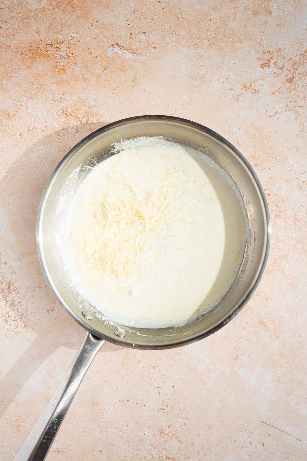 A skillet with an alfredo sauce base topped with shredded parmesan cheese. The skillet is on a tan counter.