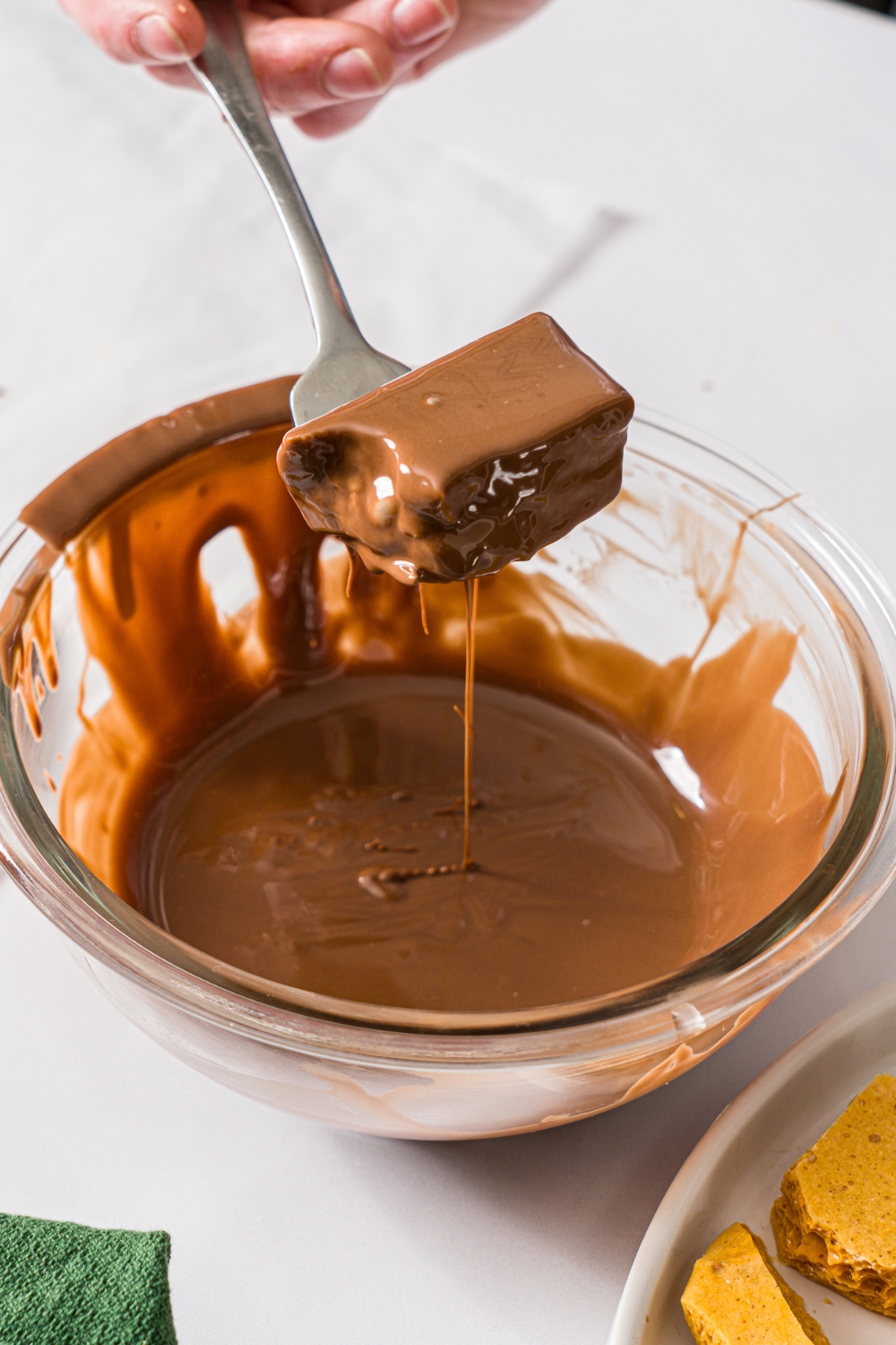 A glass bowl with melted chocolate with a fork dipping a piece of sponge candy into the chocolate. The bowl is on a marble counter.