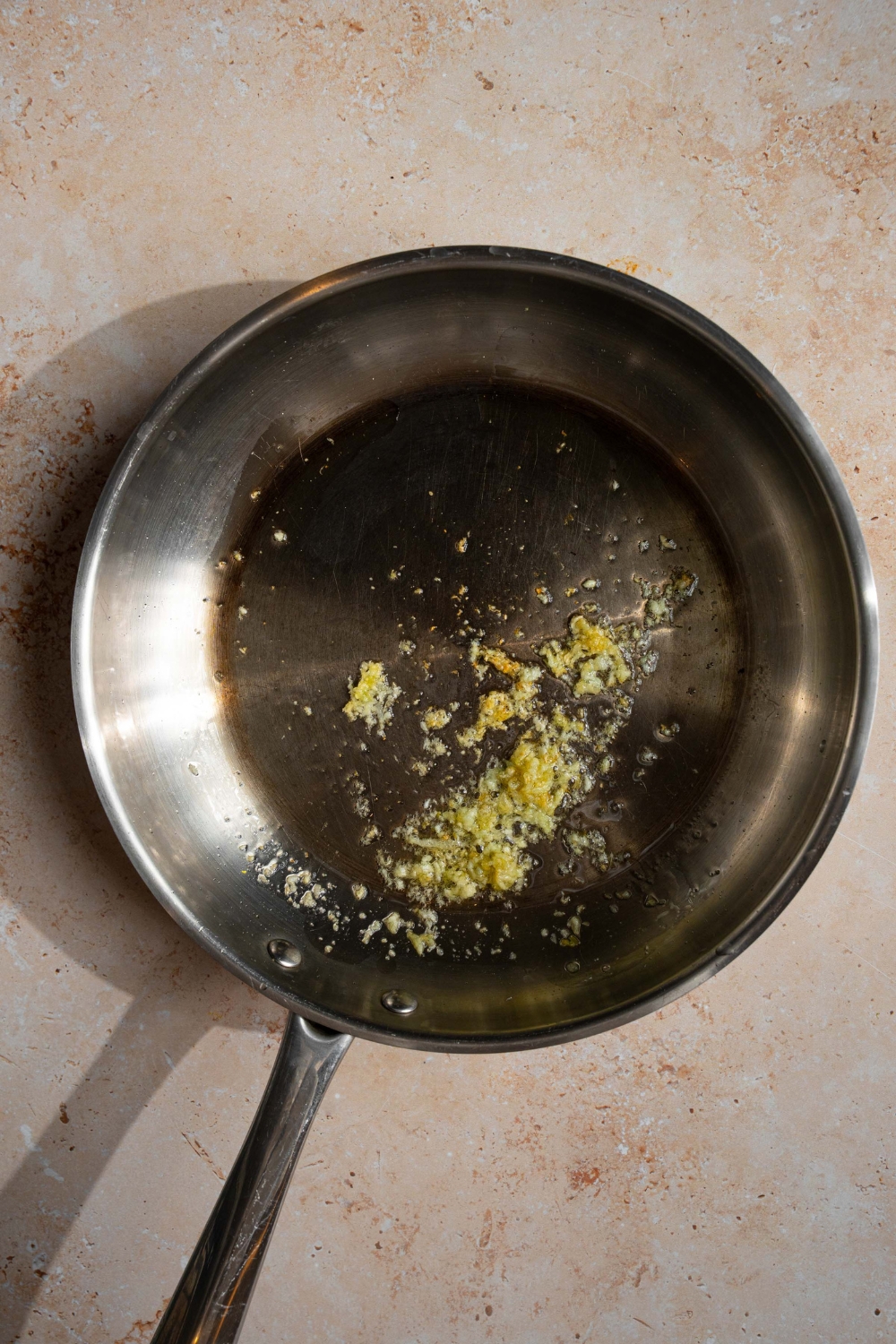 A skillet with garlic sautéing in olive oil. The skillet is on a tan counter.