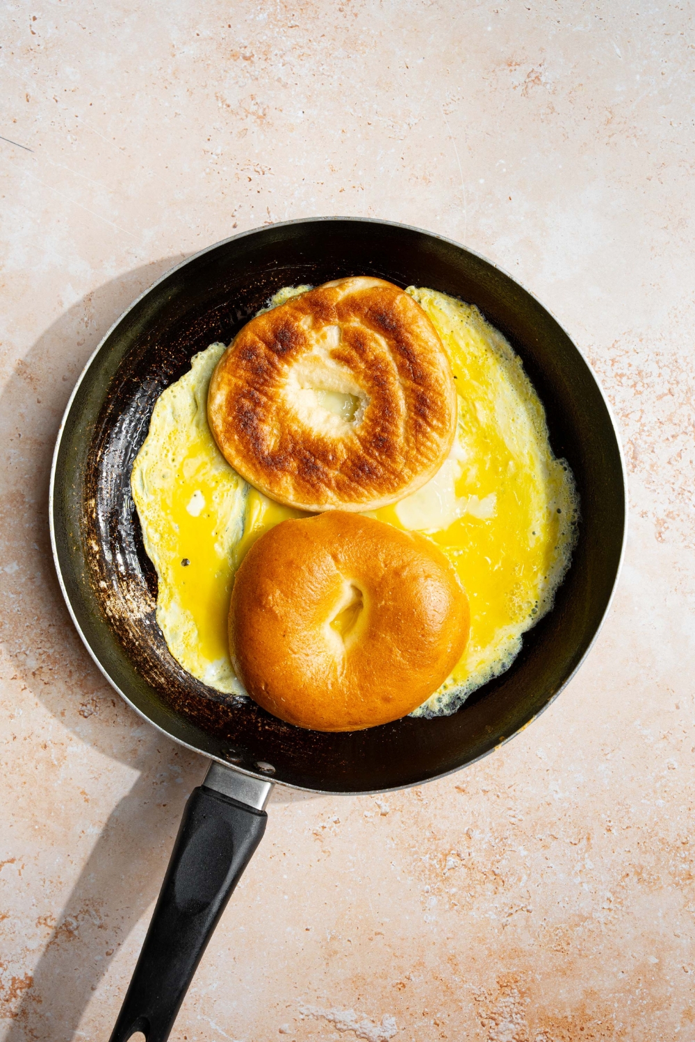 A skillet with scrambled eggs with a sliced bagel faced down on the eggs. The skillet is on a tan counter.