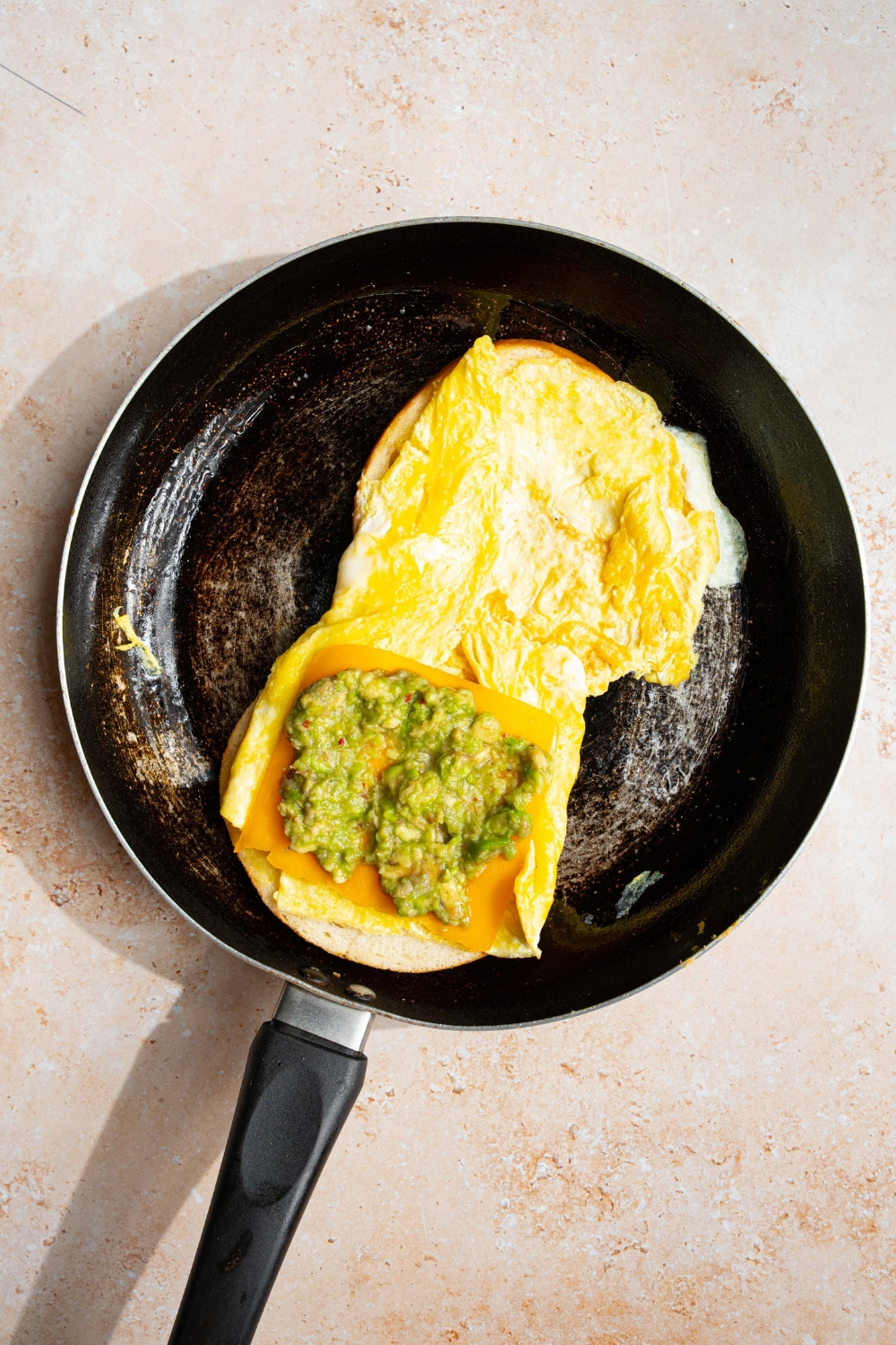 A skillet with an open bagel sandwich topped with scrambled eggs, cheese, and mashed avocado. The skillet is on a tan counter.