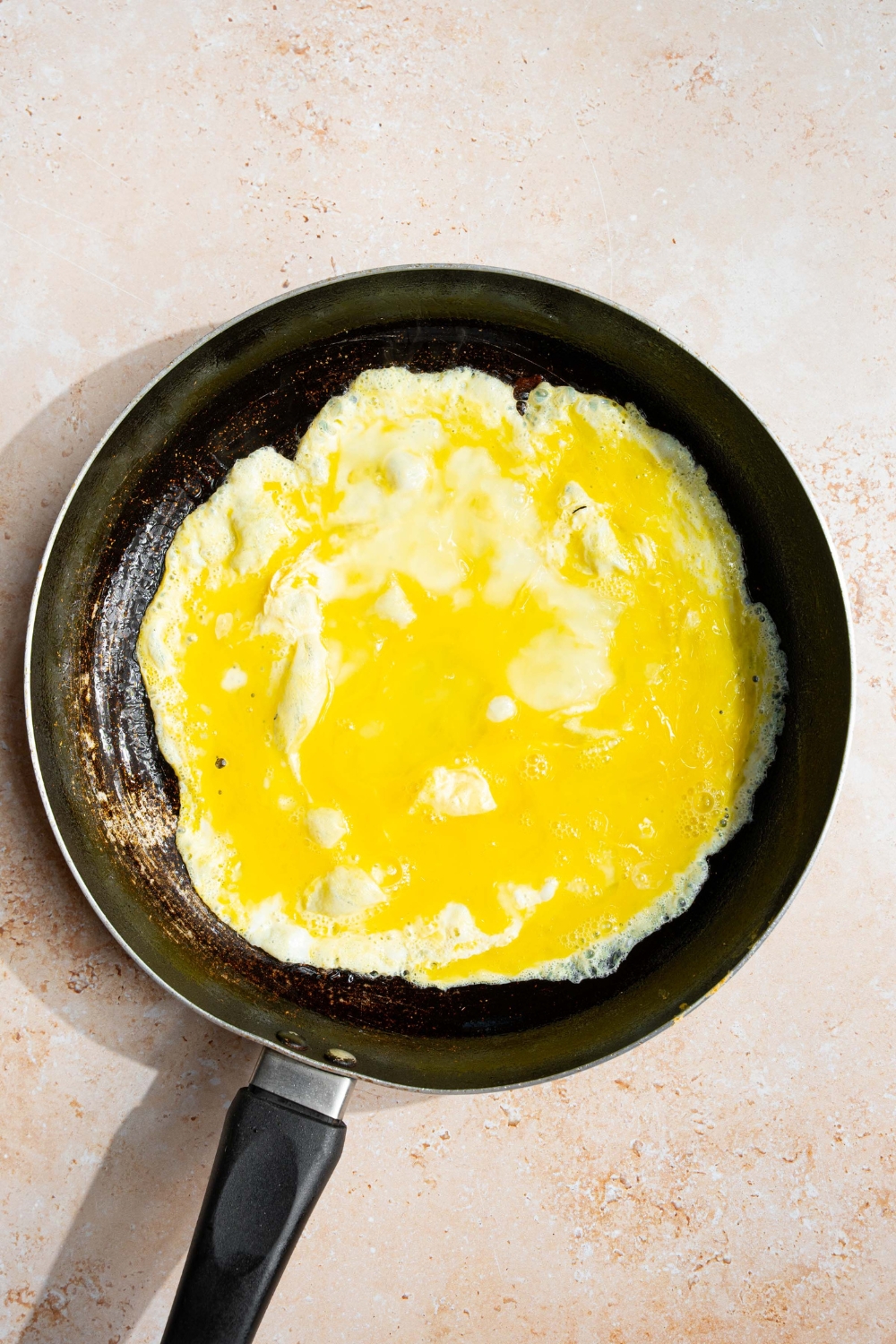 A skillet with scrambled eggs cooking in butter. The skillet is on a tan counter.