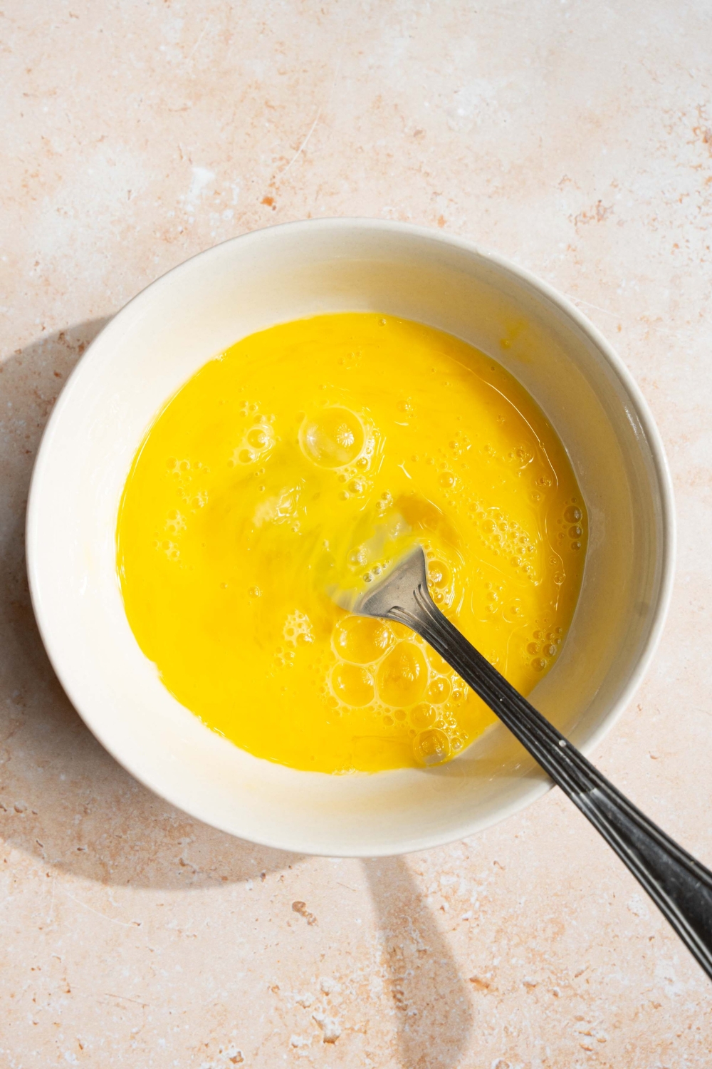 A white bowl with a fork whisking eggs. The bowl is on a tan counter.