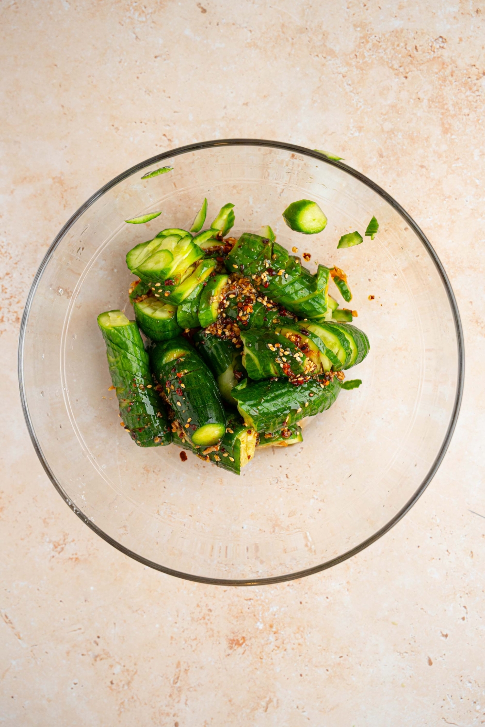 A glass bowl with spiraled cucumbers topped with spicy Asian dressing. The bowl is on a tan counter.
