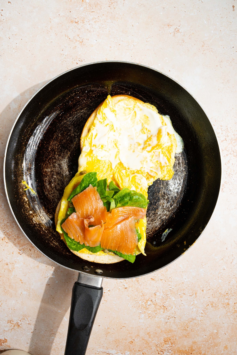 A skillet with an open loaded bagel sandwich topped with cheese, mashed avocado, spinach, and smoked salmon. The skillet is on a tan counter.