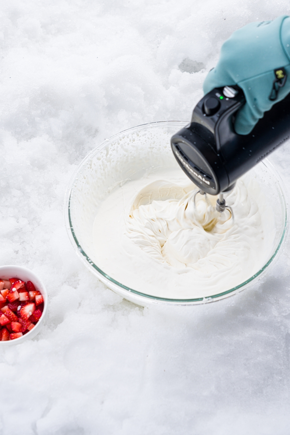 A glass bowl in a pile of snow with a hand mixer mixing ice cream ingredients. There is a small bowl of diced strawberries next to the bowl.