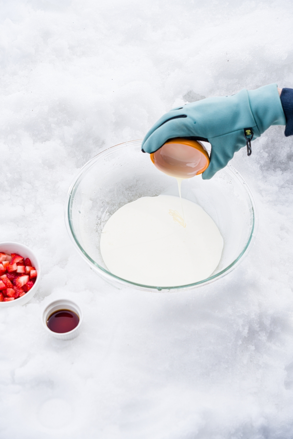 A large glass bowl sitting in snow with a hand pouring ingredients to make ice cream including heavy cream and sweetened condensed milk. The bowl is next to small bowl of strawberries and small bowl of vanilla.