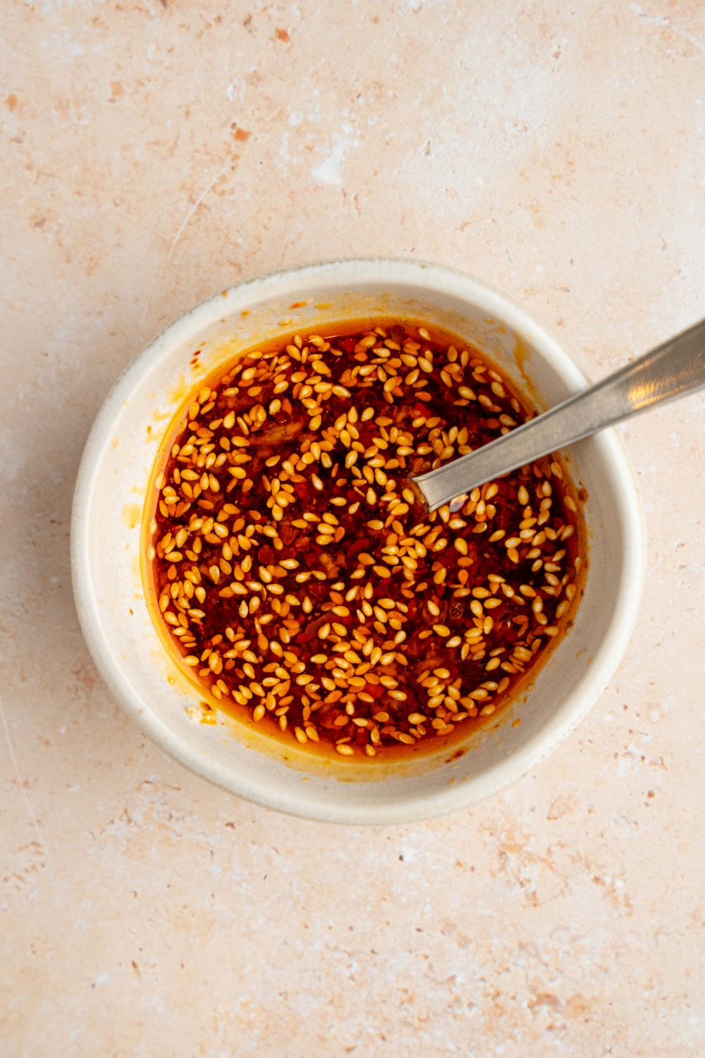A small white bowl with a spoon mixing ingredients to make spicy Asian dressing including soy sauce, sesame seeds, and chili oil. The bowl is on a tan counter.