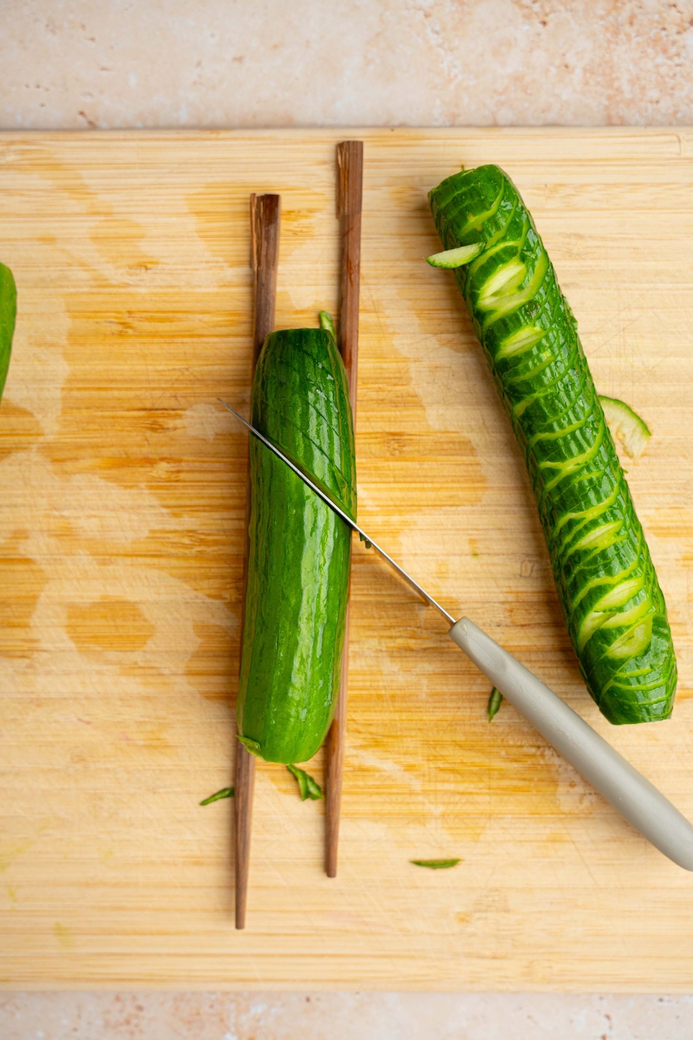 A wooden cutting board with a mini cucumber in between two chopsticks with a knife slicing angled cuts into the cucumber. The board is on a tan counter.