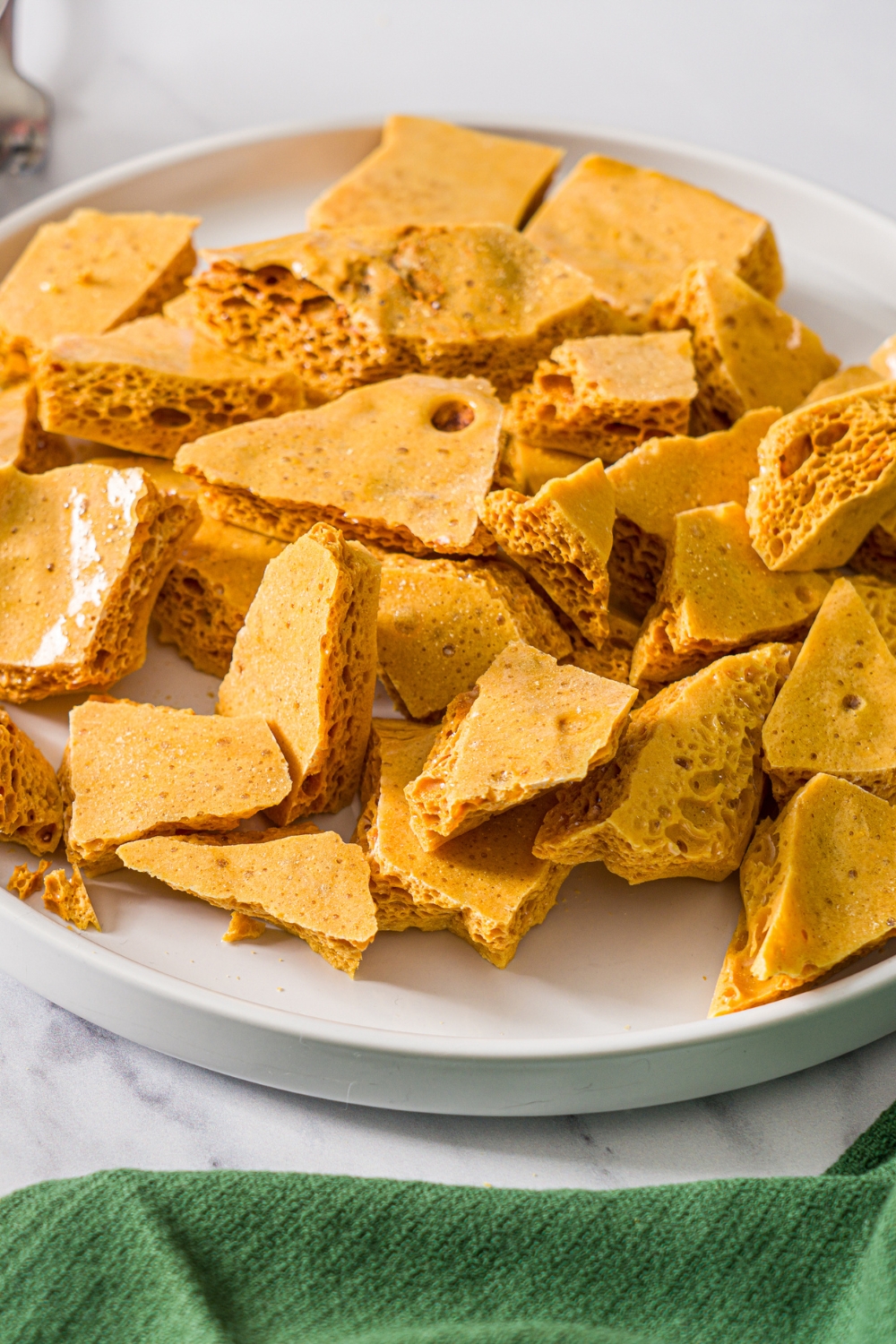 A white plate with broken pieces of sponge candy without chocolate. The plate is on a marble counter with a green cloth napkin.