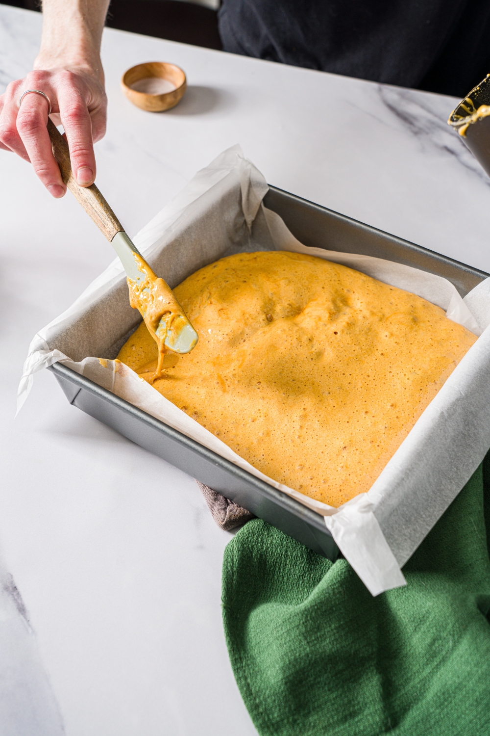 A square baking pan lined with parchment paper with a knife spreading sponge candy mixture in the pan. The pan is on a marble counter with a marble counter and a green cloth napkin.