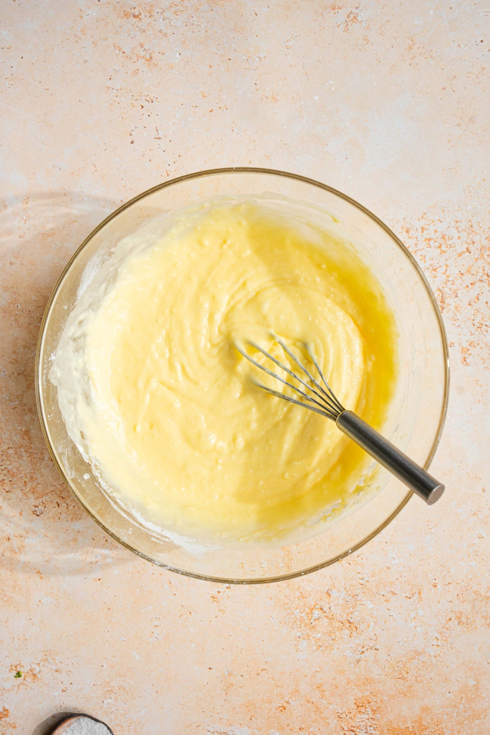 A glass bowl with a whisk mixing vanilla muffin batter. The bowl is on a tan counter.