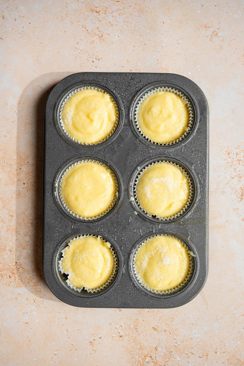 A muffin tin with unbaked vanilla muffins. The tin is on a tan counter.