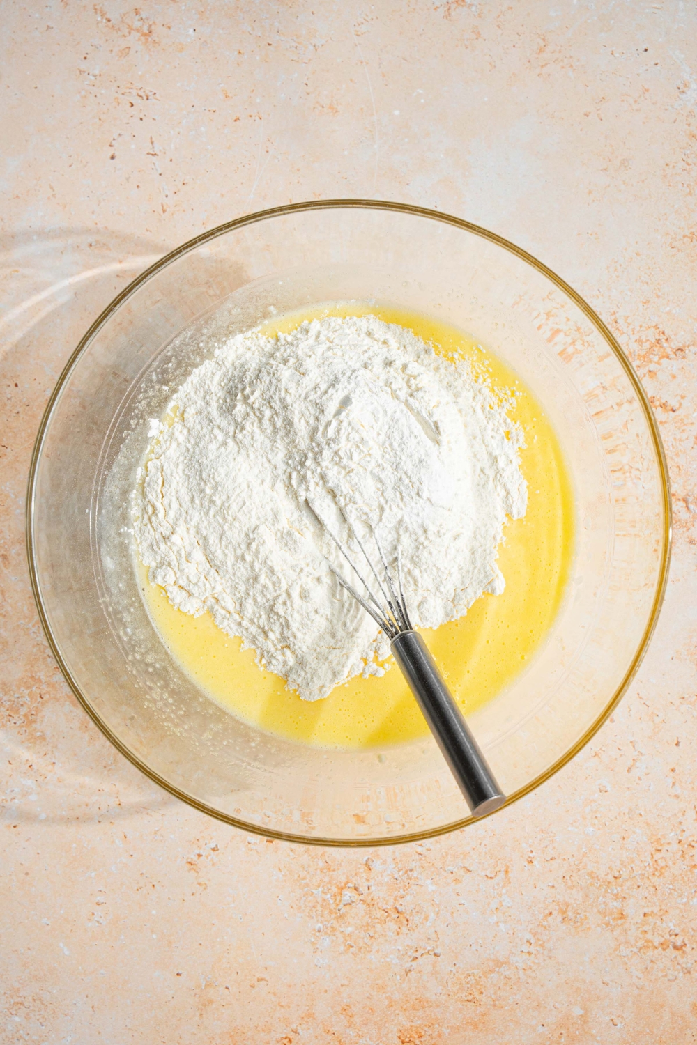 A glass bowl with a whisk combining dry ingredients with wet ingredients for vanilla muffins. The bowl is on a tan counter.