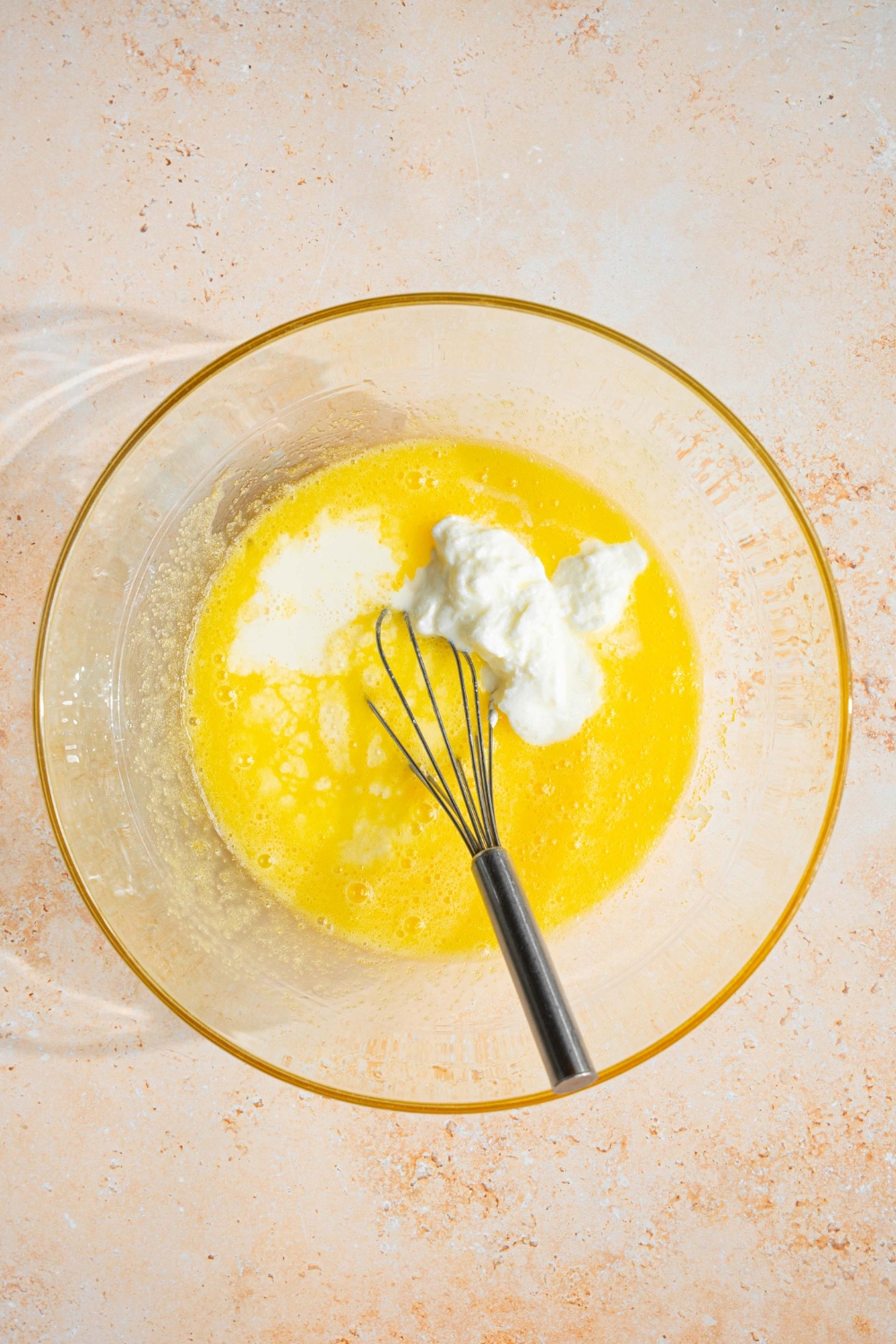 A glass bowl with a whisk mixing a butter sugar mixture with yogurt and heavy cream. The bowl is on a tan counter.