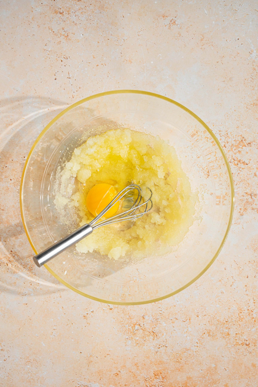 A glass bowl with a whisk mixing sugar and eggs. The bowl is on a tan counter.