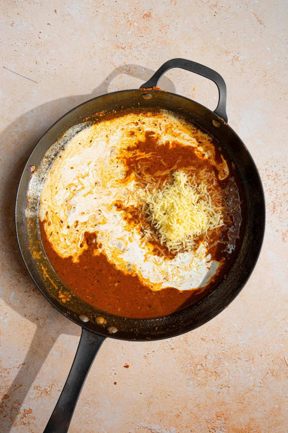 A skillet with sautéed onion, garlic, and flour with chicken broth, heavy cream, and shredded parmesan. The skillet is on a tan counter.