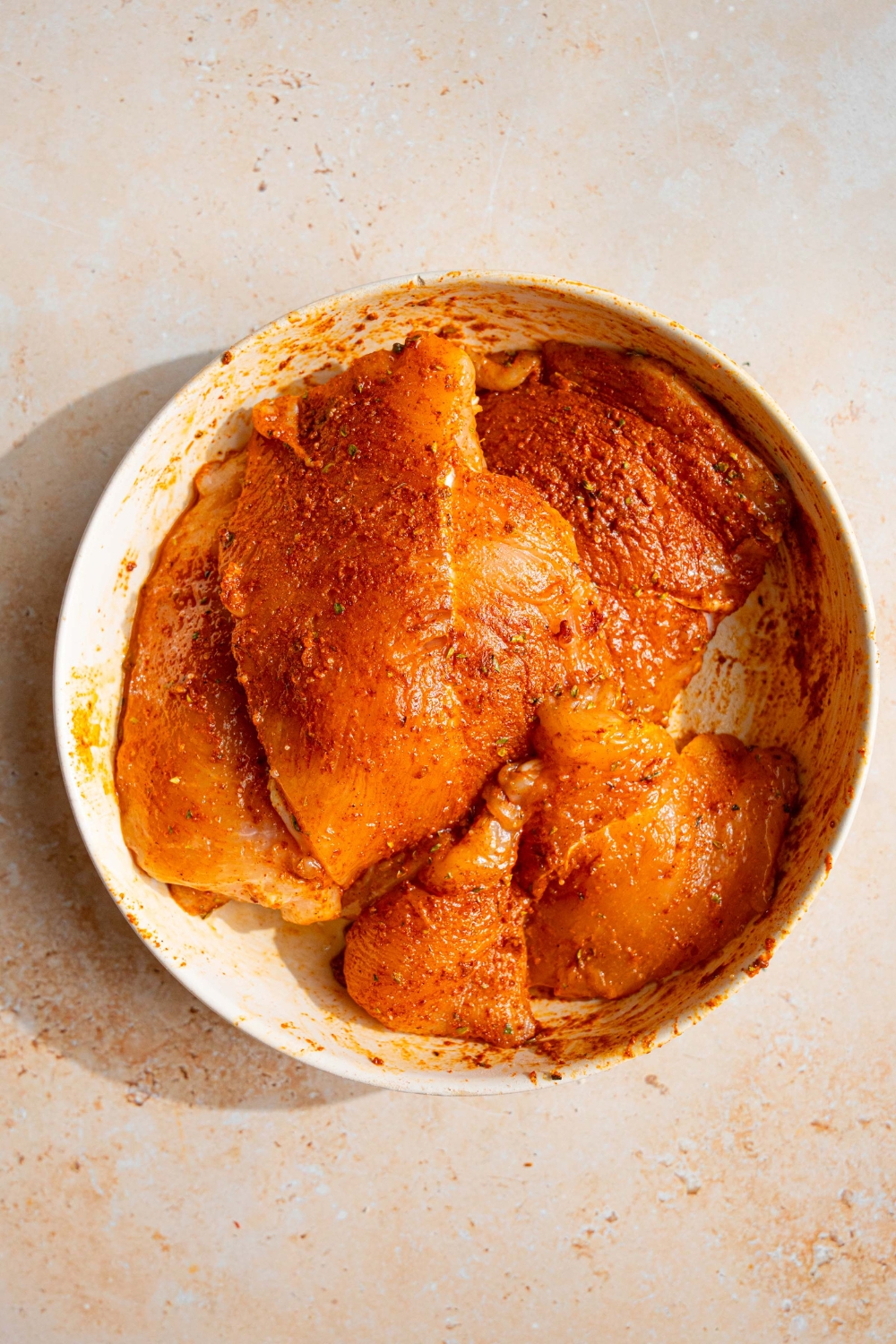 A white bowl with pounded chicken breasts tossed in a seasonings mixture. The bowl is on a tan counter.