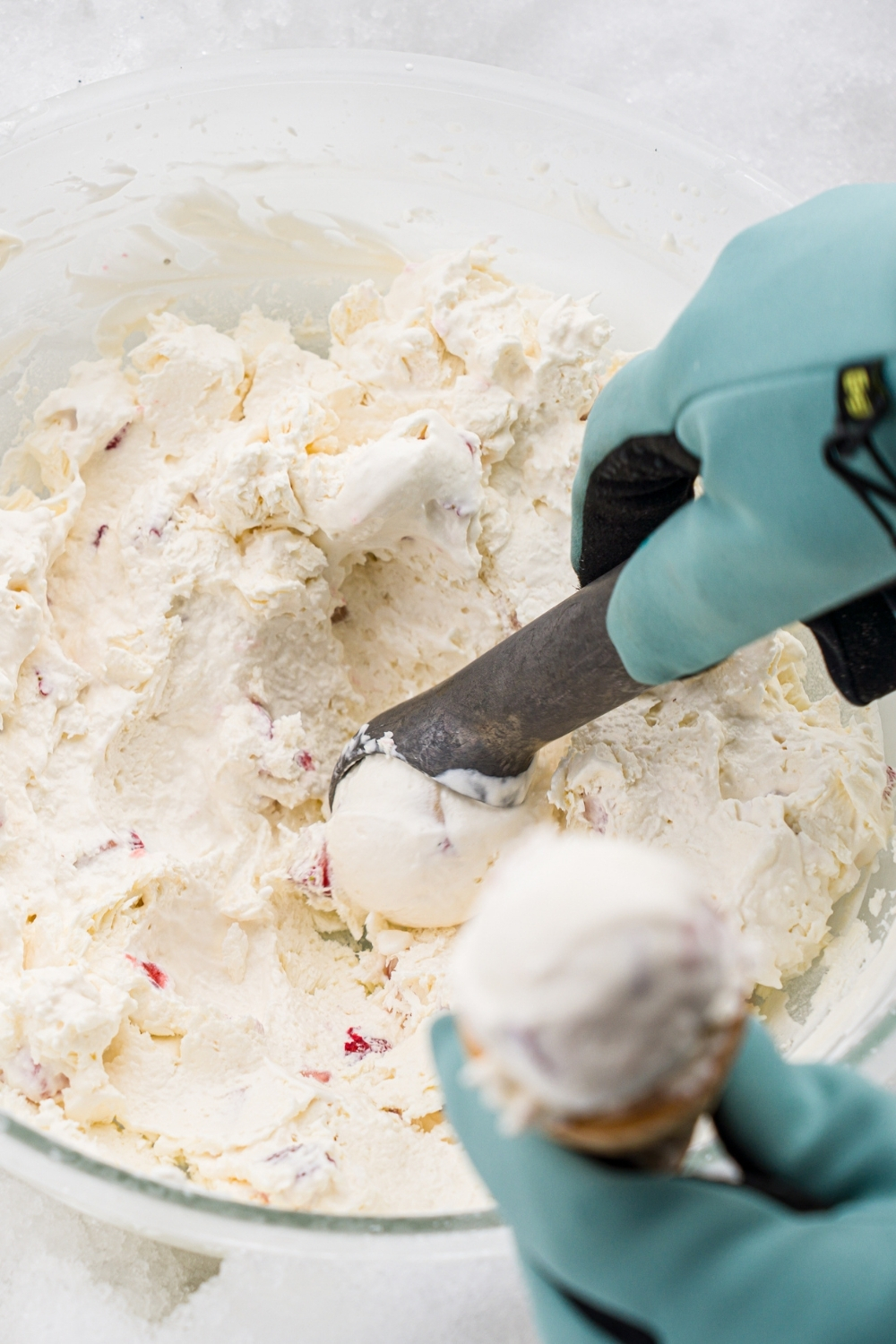 A glass bowl with hardened ice cream sitting in snow. A hand is using an ice cream scoop to scoop ice cream onto a cone.