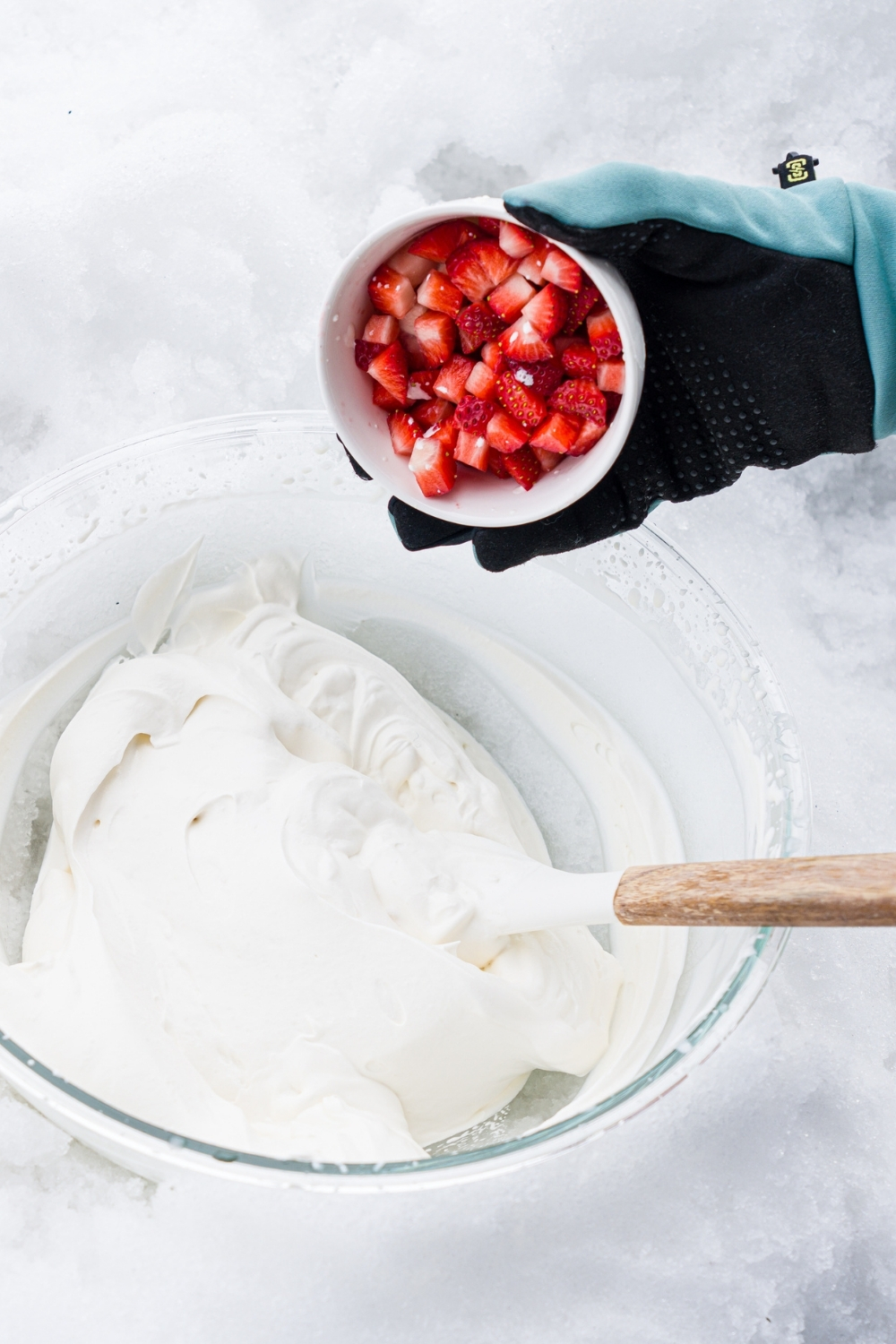 A glass bowl in snow with a whisk mixing ice cream ingredients. A small bowl of strawberries is being added to the mixture.