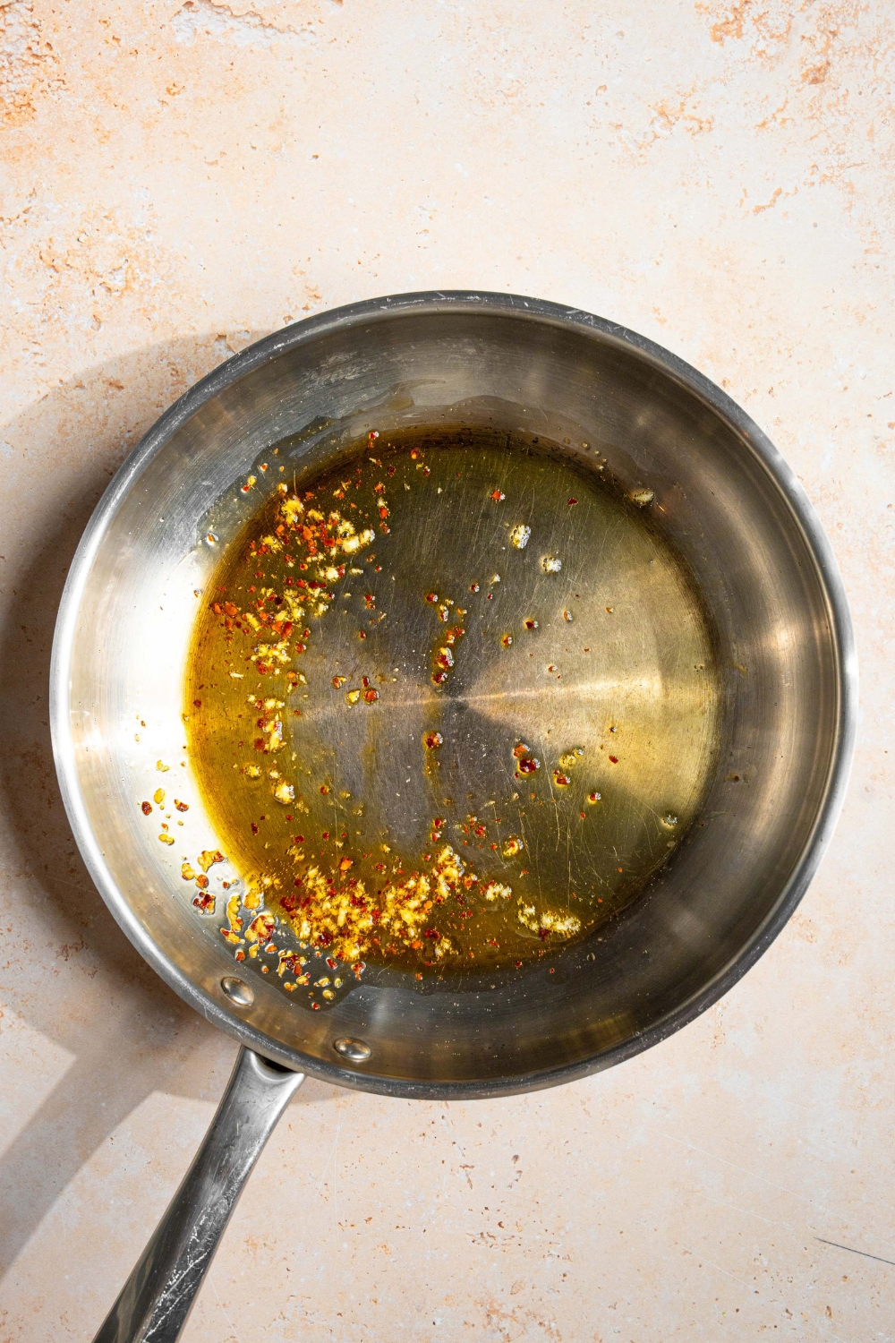 A skillet with crushed red pepper and garlic sautéing in oil. The skillet is on a tan counter.