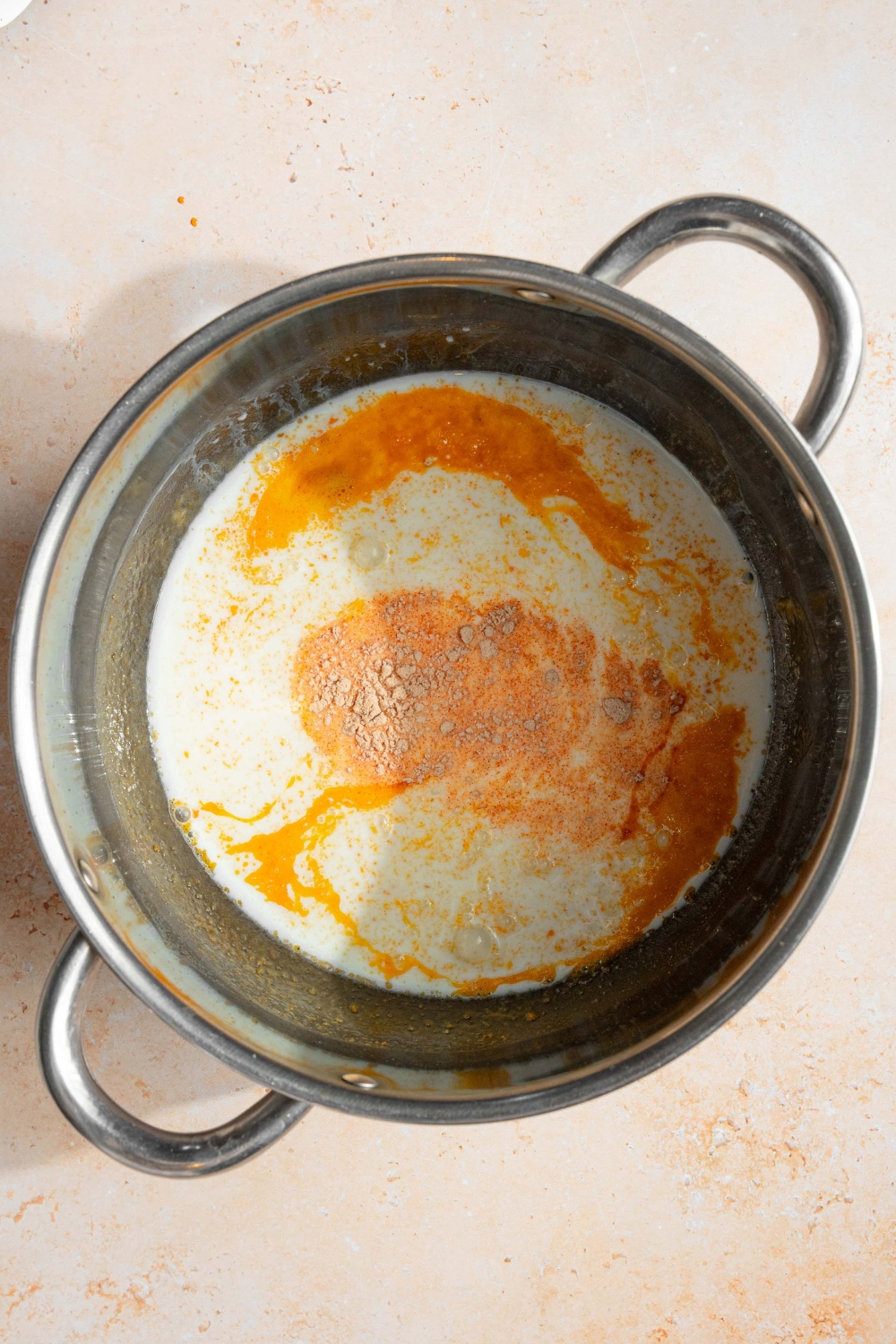 A pot with ingredients to make three cheese sauce including evaporated milk, heavy cream, and seasonings. The pot is on a tan counter.