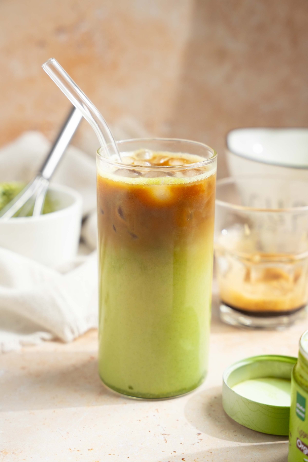 A tall glass of iced matcha latte with a glass straw. The glass is on a tan counter with a tin of matcha powder, small glass of poured espresso, and a small bowl and whisk with matcha.